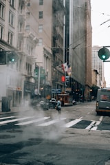 A city street scene featuring tall buildings with storefronts and fire escapes. Steam rises from a manhole cover in the foreground, while a person crosses the street. A bus and a classic cable car are visible on the road. Flags adorn one of the buildings, and the streetlights are green.