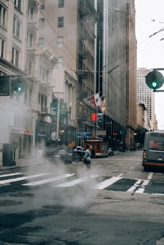 A city street scene featuring tall buildings with storefronts and fire escapes. Steam rises from a manhole cover in the foreground, while a person crosses the street. A bus and a classic cable car are visible on the road. Flags adorn one of the buildings, and the streetlights are green.