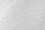 A close-up view of a textured white surface, possibly a plastered or stucco wall. The surface has a rough, uneven finish with small variations and grooves, creating a subtle pattern.