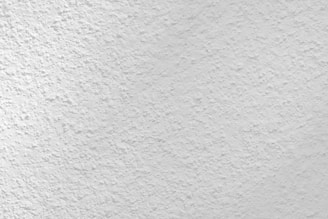 A close-up view of a textured white surface, possibly a plastered or stucco wall. The surface has a rough, uneven finish with small variations and grooves, creating a subtle pattern.