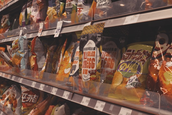 A store shelf filled with various packages of snacks, including colorful bags of chips with different designs and labels. Several bags feature text in different languages, including English and what appears to be Chinese or Korean. Price tags are visible on the shelf.