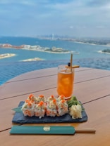A plate of sushi rolls garnished with orange roe, accompanied by wasabi and pickled ginger, rest on a slate serving board atop a wooden table. Next to it is a tall glass of iced tea with a garnish. The background features a stunning panoramic view of a coastal city and a vast body of water with scattered islands under a partly cloudy sky.