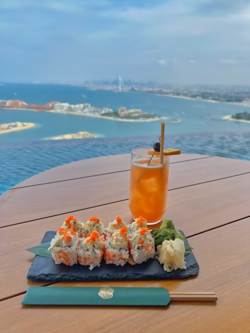 A plate of sushi rolls garnished with orange roe, accompanied by wasabi and pickled ginger, rest on a slate serving board atop a wooden table. Next to it is a tall glass of iced tea with a garnish. The background features a stunning panoramic view of a coastal city and a vast body of water with scattered islands under a partly cloudy sky.
