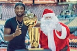 A man in a black shirt stands next to a person dressed as Santa Claus, both smiling and gesturing positively with a large, gold trophy between them. The setting appears to be an indoor stadium with rows of empty seats in the background.