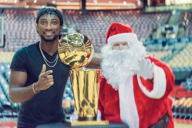 A man in a black shirt stands next to a person dressed as Santa Claus, both smiling and gesturing positively with a large, gold trophy between them. The setting appears to be an indoor stadium with rows of empty seats in the background.