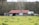 A rustic barn with a red roof sits in a grassy field, surrounded by wooden fences and trees. There are metal tanks positioned beside the barn.