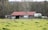 A rustic barn with a red roof sits in a grassy field, surrounded by wooden fences and trees. There are metal tanks positioned beside the barn.