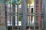 A wooden fence with a grid-like pattern is set against a backdrop of brick and greenery. The fence is weathered, with a natural, rustic appearance. In the background, brick structures and blurred foliage create a layered, textured scene.