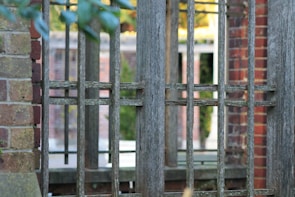 A wooden fence with a grid-like pattern is set against a backdrop of brick and greenery. The fence is weathered, with a natural, rustic appearance. In the background, brick structures and blurred foliage create a layered, textured scene.