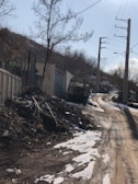 A narrow dirt road is lined with snow and tracked by vehicle tire marks. There is a utility vehicle parked by the side of the road near a fence. The area is bordered by trees with barren branches, suggesting a winter setting. Telephone poles with wires run alongside the road.