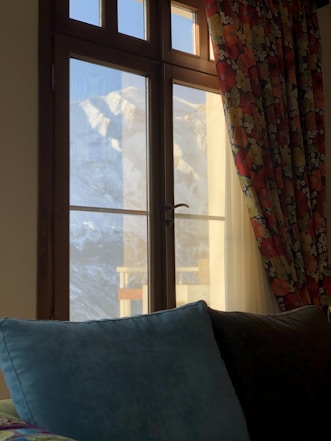 A large window with wooden frames overlooks a mountain view with snow-covered peaks. The room interior features a vibrant multi-colored floral curtain on one side and two soft pillows, one in a deep teal hue, placed on a dark surface.