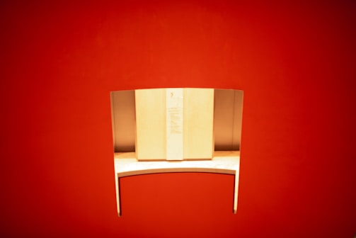 A sleek, minimalist wooden desk with a curved metal privacy divider is set against a vibrant red background. The desk is well-lit, highlighting its smooth surface and modern design.