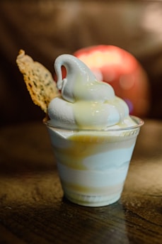 A creamy soft-serve ice cream in a clear plastic cup drizzled with a light yellow sauce. A piece of thin, crispy wafer is placed on the side. The ice cream has a smooth, swirled texture and is set against a blurred, warm-toned background.