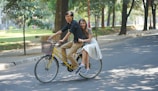 A couple rides on a bicycle along a tree-lined street, with the woman sitting on the back while holding onto the man who is pedaling. The street is shaded by trees, creating a pleasant and relaxed atmosphere.