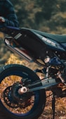 A close-up view of a motorcycle, focusing on the rear wheel and exhaust area. The bike is parked on a grassy or earthy surface, and a person wearing a plaid shirt is partially visible, holding onto the handlebar. The motorcycle has a sporty design with labels such as 'Dirty Heroes' and '225' visible on the body.