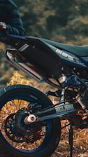 A close-up view of a motorcycle, focusing on the rear wheel and exhaust area. The bike is parked on a grassy or earthy surface, and a person wearing a plaid shirt is partially visible, holding onto the handlebar. The motorcycle has a sporty design with labels such as 'Dirty Heroes' and '225' visible on the body.