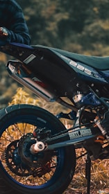 A close-up view of a motorcycle, focusing on the rear wheel and exhaust area. The bike is parked on a grassy or earthy surface, and a person wearing a plaid shirt is partially visible, holding onto the handlebar. The motorcycle has a sporty design with labels such as 'Dirty Heroes' and '225' visible on the body.