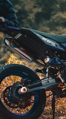A close-up view of a motorcycle, focusing on the rear wheel and exhaust area. The bike is parked on a grassy or earthy surface, and a person wearing a plaid shirt is partially visible, holding onto the handlebar. The motorcycle has a sporty design with labels such as 'Dirty Heroes' and '225' visible on the body.