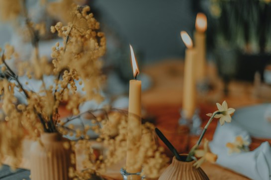 A warm, softly lit scene featuring several lit candles with small flames flickering. The setting includes delicate floral arrangements in muted yellow tones, set against a blurred background with hints of tableware. The overall composition creates a soothing and intimate atmosphere.