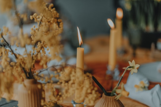 A warm, softly lit scene featuring several lit candles with small flames flickering. The setting includes delicate floral arrangements in muted yellow tones, set against a blurred background with hints of tableware. The overall composition creates a soothing and intimate atmosphere.