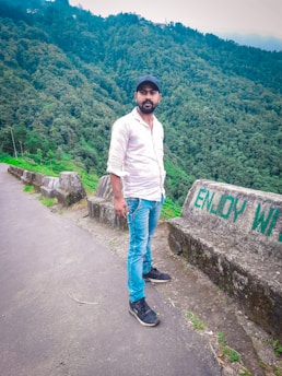 A man stands on a paved pathway next to a barrier with the words 'ENJOY WILD.' He is wearing a white shirt, blue jeans, and a black cap. The background is filled with lush, green forested hills, suggesting a scenic and natural setting.