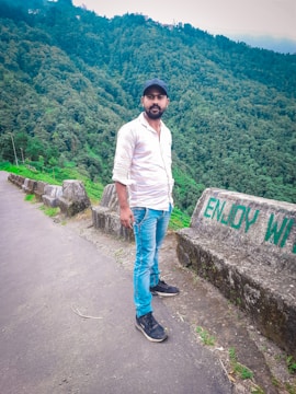 A man stands on a paved pathway next to a barrier with the words 'ENJOY WILD.' He is wearing a white shirt, blue jeans, and a black cap. The background is filled with lush, green forested hills, suggesting a scenic and natural setting.