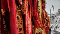A collection of vibrant red and gold woven fabrics featuring intricate designs and symbols. These textiles hang in vertical rows, creating a rich tapestry-like effect. To the right, a vintage street lamp stands on a stone path, with trees in the background, suggesting an outdoor setting.