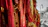 A collection of vibrant red and gold woven fabrics featuring intricate designs and symbols. These textiles hang in vertical rows, creating a rich tapestry-like effect. To the right, a vintage street lamp stands on a stone path, with trees in the background, suggesting an outdoor setting.