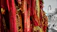 A collection of vibrant red and gold woven fabrics featuring intricate designs and symbols. These textiles hang in vertical rows, creating a rich tapestry-like effect. To the right, a vintage street lamp stands on a stone path, with trees in the background, suggesting an outdoor setting.