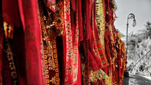 A collection of vibrant red and gold woven fabrics featuring intricate designs and symbols. These textiles hang in vertical rows, creating a rich tapestry-like effect. To the right, a vintage street lamp stands on a stone path, with trees in the background, suggesting an outdoor setting.