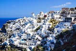 A picturesque hillside village with whitewashed buildings and bright blue domes overlooking a deep blue sea. The architecture features winding paths and terraces, with the presence of windmills adding to the scenic beauty.