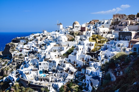 A picturesque hillside village with whitewashed buildings and bright blue domes overlooking a deep blue sea. The architecture features winding paths and terraces, with the presence of windmills adding to the scenic beauty.