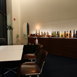 A contemporary dining space featuring a row of assorted bottles and decor items on a sleek wooden countertop. Wooden chairs are arranged around a table with a contrasting black and white design. A potted plant and an oval mirror add decorative elements to the minimalist interior.
