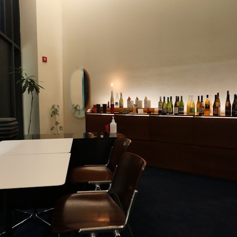A contemporary dining space featuring a row of assorted bottles and decor items on a sleek wooden countertop. Wooden chairs are arranged around a table with a contrasting black and white design. A potted plant and an oval mirror add decorative elements to the minimalist interior.