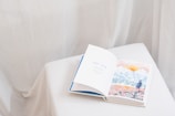 An open book rests on a white, cloth-covered surface. The left page displays the text 'part one' in blue letters, while the right page features a watercolor illustration of a landscape with mountains, a sun, and a person in a hat looking at the scenery. The background is soft and neutral, with draped fabric creating a gentle ambiance.