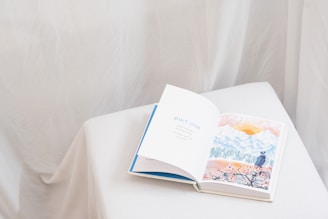 An open book rests on a white, cloth-covered surface. The left page displays the text 'part one' in blue letters, while the right page features a watercolor illustration of a landscape with mountains, a sun, and a person in a hat looking at the scenery. The background is soft and neutral, with draped fabric creating a gentle ambiance.