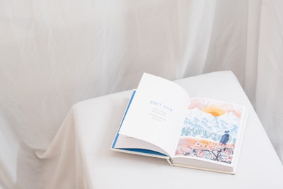 An open book rests on a white, cloth-covered surface. The left page displays the text 'part one' in blue letters, while the right page features a watercolor illustration of a landscape with mountains, a sun, and a person in a hat looking at the scenery. The background is soft and neutral, with draped fabric creating a gentle ambiance.