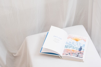 An open book rests on a white, cloth-covered surface. The left page displays the text 'part one' in blue letters, while the right page features a watercolor illustration of a landscape with mountains, a sun, and a person in a hat looking at the scenery. The background is soft and neutral, with draped fabric creating a gentle ambiance.