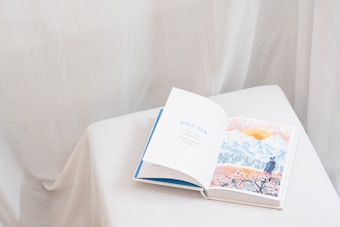 An open book rests on a white, cloth-covered surface. The left page displays the text 'part one' in blue letters, while the right page features a watercolor illustration of a landscape with mountains, a sun, and a person in a hat looking at the scenery. The background is soft and neutral, with draped fabric creating a gentle ambiance.