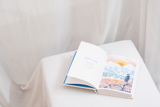 An open book rests on a white, cloth-covered surface. The left page displays the text 'part one' in blue letters, while the right page features a watercolor illustration of a landscape with mountains, a sun, and a person in a hat looking at the scenery. The background is soft and neutral, with draped fabric creating a gentle ambiance.