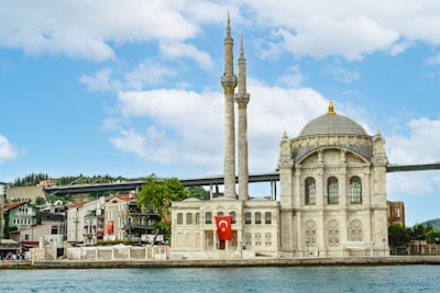 A grand mosque with two tall minarets stands next to a waterway. The architecture is intricate, with ornate detailing and a large dome. A red Turkish flag is prominently displayed on the building. In the background, a bridge spans across, accompanied by several trees and small houses lined along the shore under a partly cloudy sky.