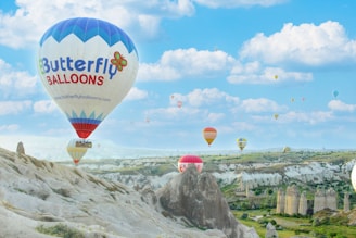 Hot air balloons of various colors and designs float over a rocky landscape with unique geological formations. A clear blue sky with scattered white clouds provides a picturesque backdrop.