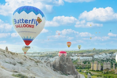 Hot air balloons of various colors and designs float over a rocky landscape with unique geological formations. A clear blue sky with scattered white clouds provides a picturesque backdrop.
