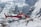 A red and white helicopter hovers above a snowy mountainous landscape, surrounded by rugged ice formations and steep cliffs.
