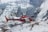 A red and white helicopter hovers above a snowy mountainous landscape, surrounded by rugged ice formations and steep cliffs.