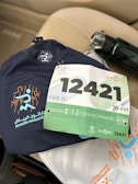 A navy blue athletic shirt with 'Riyadh Marathon' and a stylized runner graphic is placed on a beige car seat. A race bib numbered 12421 with green and white colors, indicating a 10 KM event scheduled for February 11, is positioned on top of the shirt. There is a water bottle with a black cap next to the shirt.