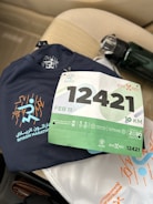 A navy blue athletic shirt with 'Riyadh Marathon' and a stylized runner graphic is placed on a beige car seat. A race bib numbered 12421 with green and white colors, indicating a 10 KM event scheduled for February 11, is positioned on top of the shirt. There is a water bottle with a black cap next to the shirt.