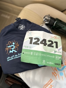 A navy blue athletic shirt with 'Riyadh Marathon' and a stylized runner graphic is placed on a beige car seat. A race bib numbered 12421 with green and white colors, indicating a 10 KM event scheduled for February 11, is positioned on top of the shirt. There is a water bottle with a black cap next to the shirt.