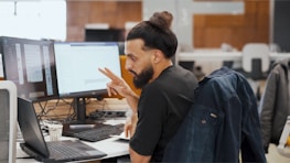 A person with long hair tied up is sitting at a desk in a modern office environment. They are looking at two computer monitors while gesturing with their hand in a thoughtful manner. A jacket is draped over the back of the chair, and a coffee cup is placed next to a laptop.