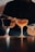 Two cocktails in stem glasses are placed on a reflective surface. One glass contains a dark beverage, while the other holds a lighter-colored drink. A person in dark attire appears in the background, using tools to prepare or garnish the drinks.